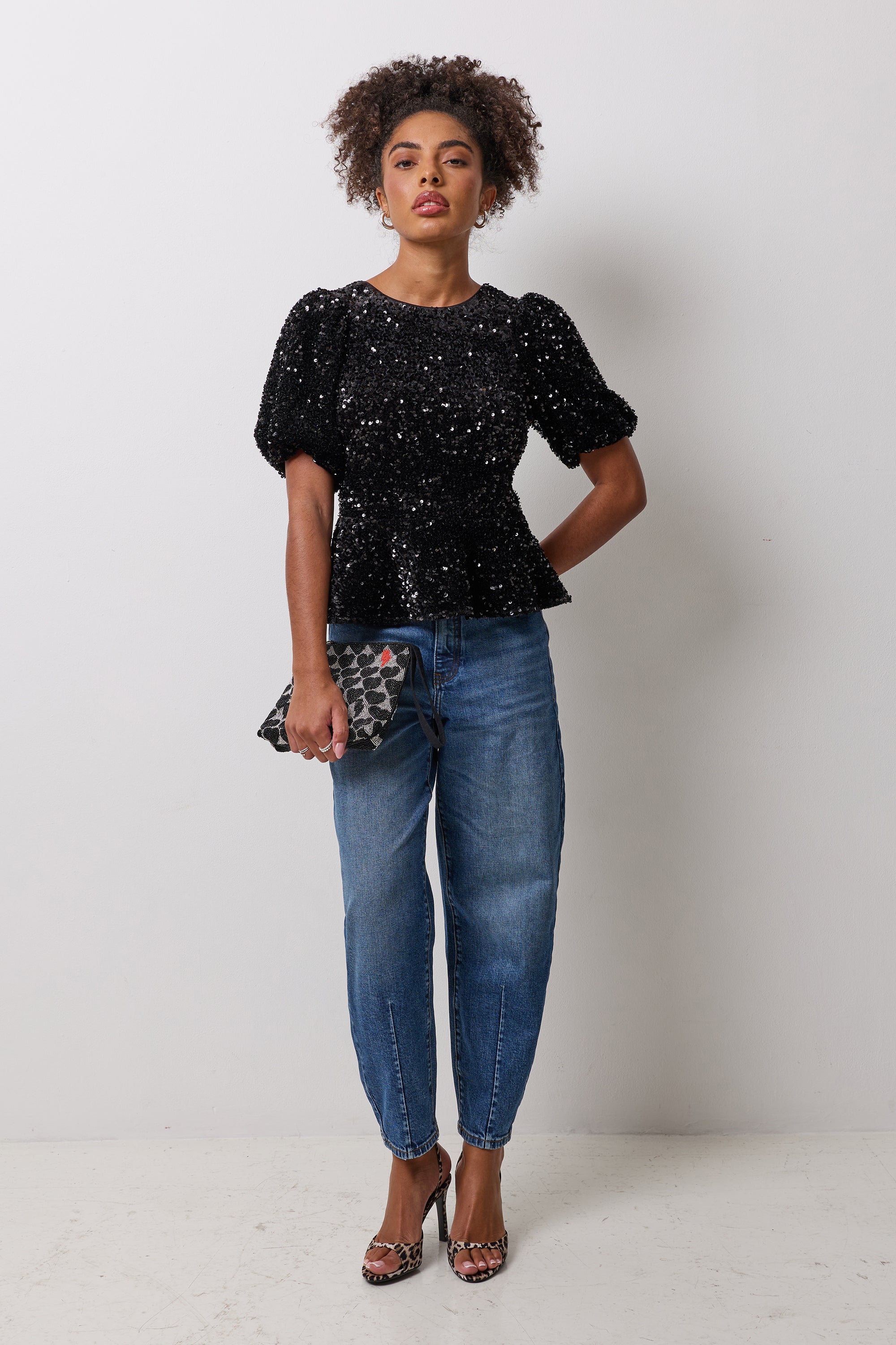 Scamp & Dude Black Sequin Peplum Top | Model wearing a black sequin peplum top with a pair of blue barrel jeans and a beaded clutch bag.