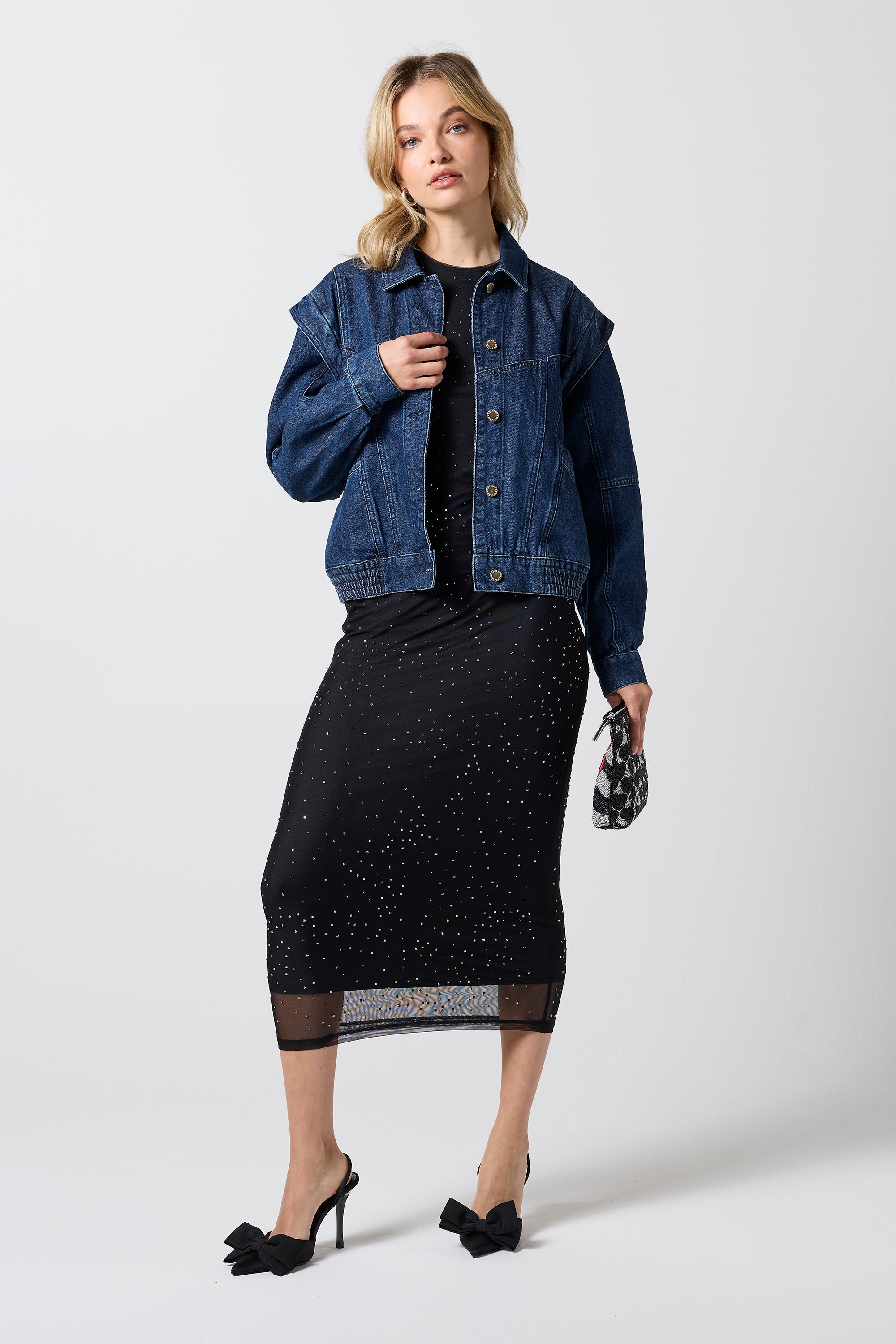 Scamp & Dude Mid Wash Seam Detail Denim Jacket | Model wearing a dark blue denim jacket over a black top, a black midi skirt with subtle silver speckles, and black pointed-toe heels. She is holding a small black-and-white beaded patterned clutch.