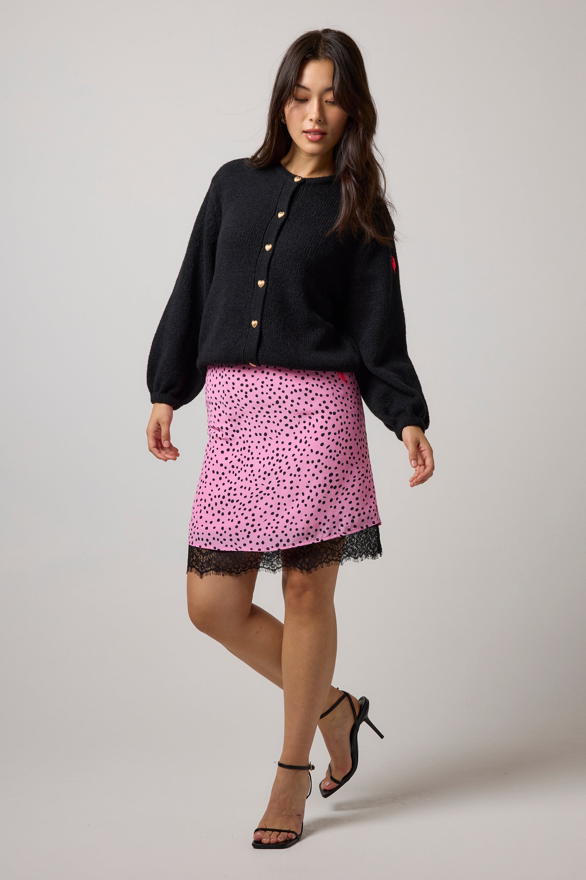 Scamp & Dude Pink With Black Cheetah Spot Lace Hem Mini Skirt | Model wearing a black cardigan with a pink cheetah print mini skirt featuring a black lace trim.