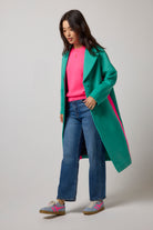 Scamp & Dude Green with Neon Pink Wool Blend Coat | Model wearing a long green coat with bright pink side panels, worn over a hot pink top with blue jeans and trainers.