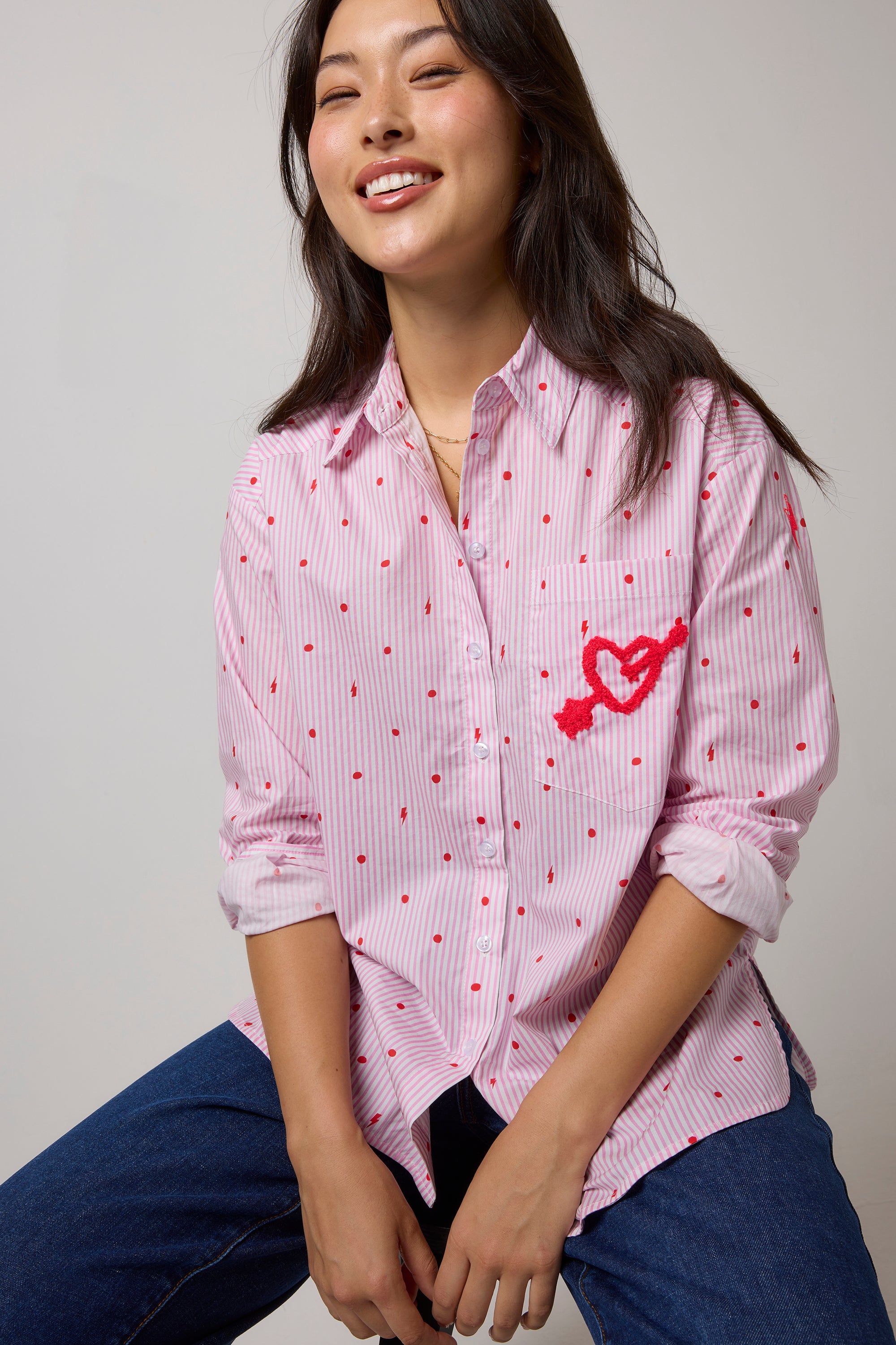 Scamp & Dude Pink Polka Dot Stripe Shirt | Model wearing a pink and white vertically striped button-up shirt with small polka dots and a red embroidered heart with arrow on the chest, paired with blue jeans.