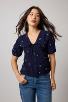 Scamp & Dude Navy Embroidered Cord Collared Blouse | Model wearing a navy blue short-sleeve top with embroidered white and red hearts and bolts worn with blue jeans.