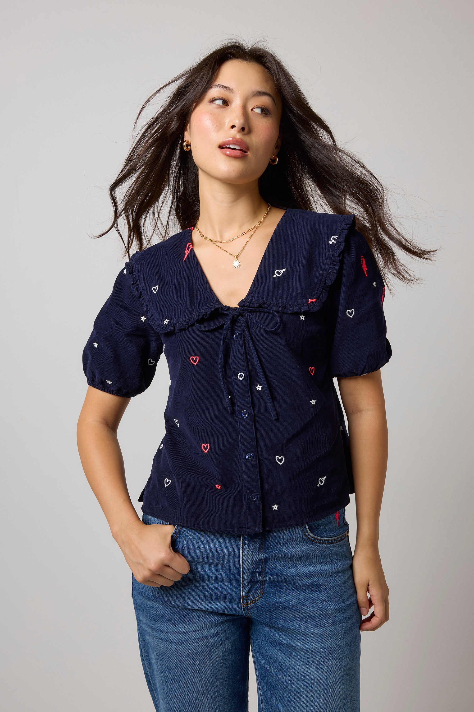 Scamp & Dude Navy Embroidered Cord Collared Blouse | Model wearing a navy blue short-sleeve top with embroidered white and red hearts and bolts worn with blue jeans.