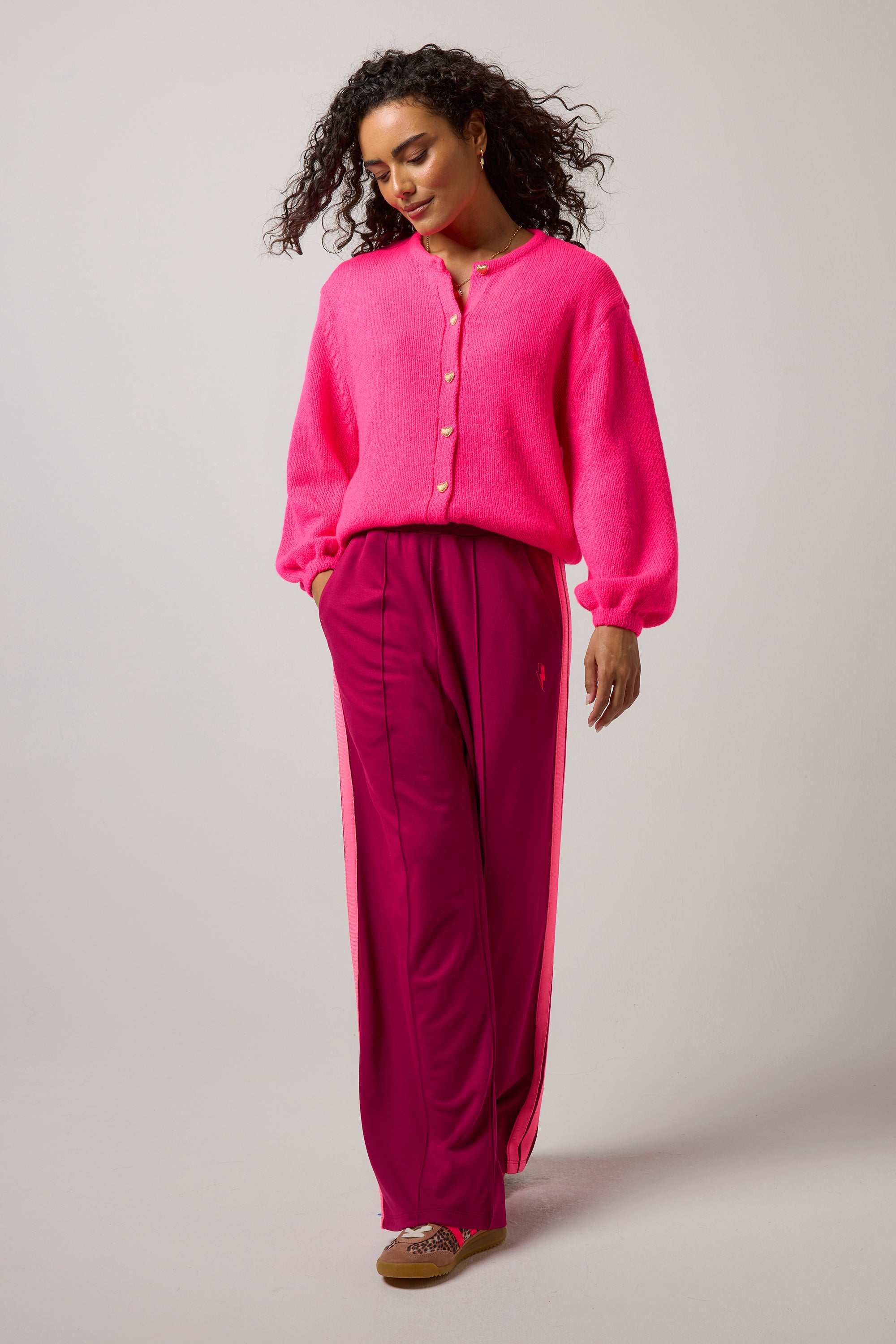 Scamp & Dude Neon Pink Cardigan | Model wearing a pair of burgundy wide leg trousers with a neon pink cardigan and animal print trainers.