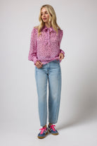 Scamp & Dude Pink with Black Cheetah Spot Tie Neck Blouse | Model wearing a pink long-sleeve blouse with a small black pattern, paired with light blue jeans and colorful sneakers.