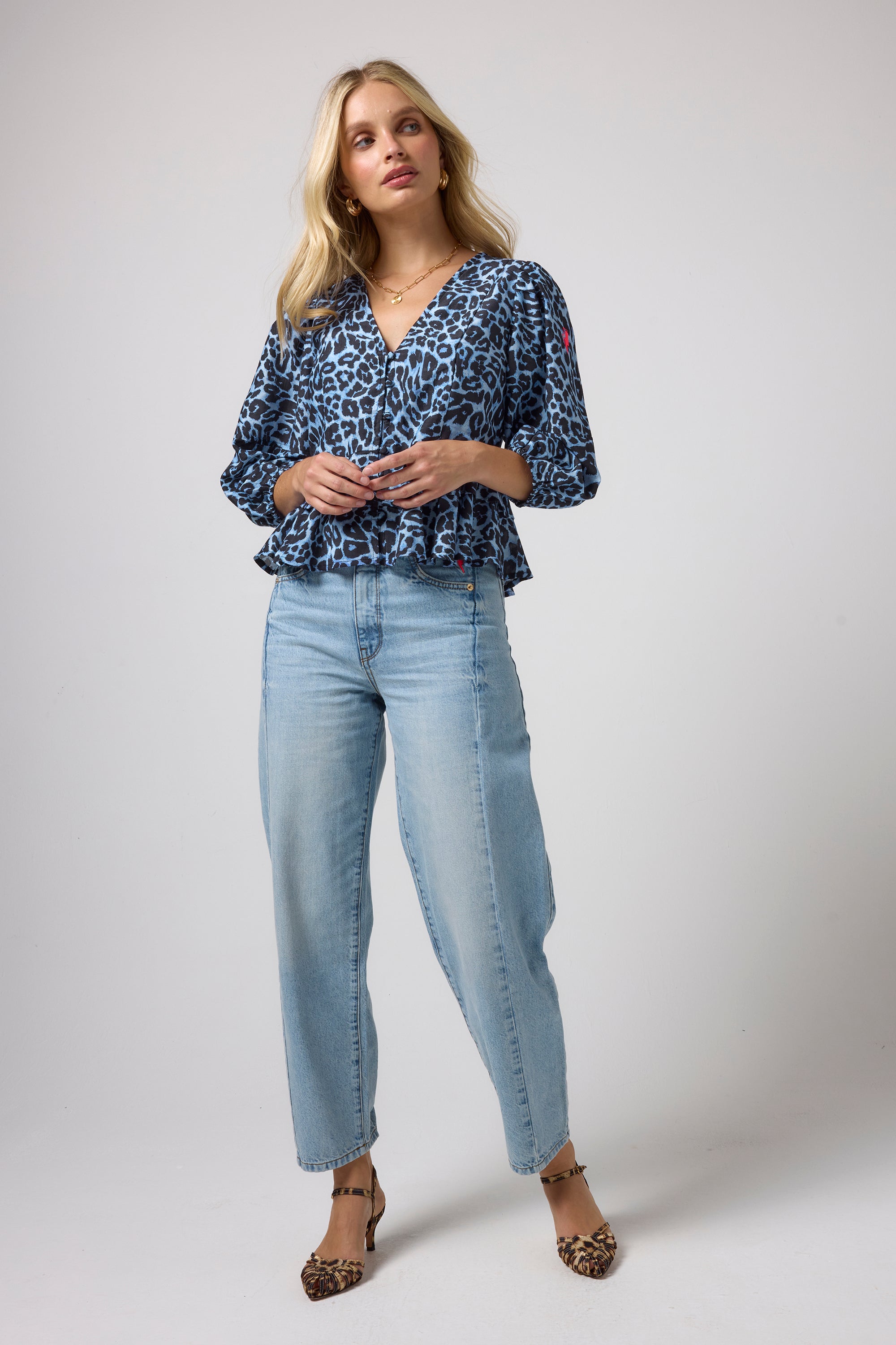 Scamp & Dude Blue Jaguar Wave Peplum Blouse | Model wearing a blue and black jaguar print blouse with three-quarter sleeves, paired with light-wash blue jeans and leopard-print heels.