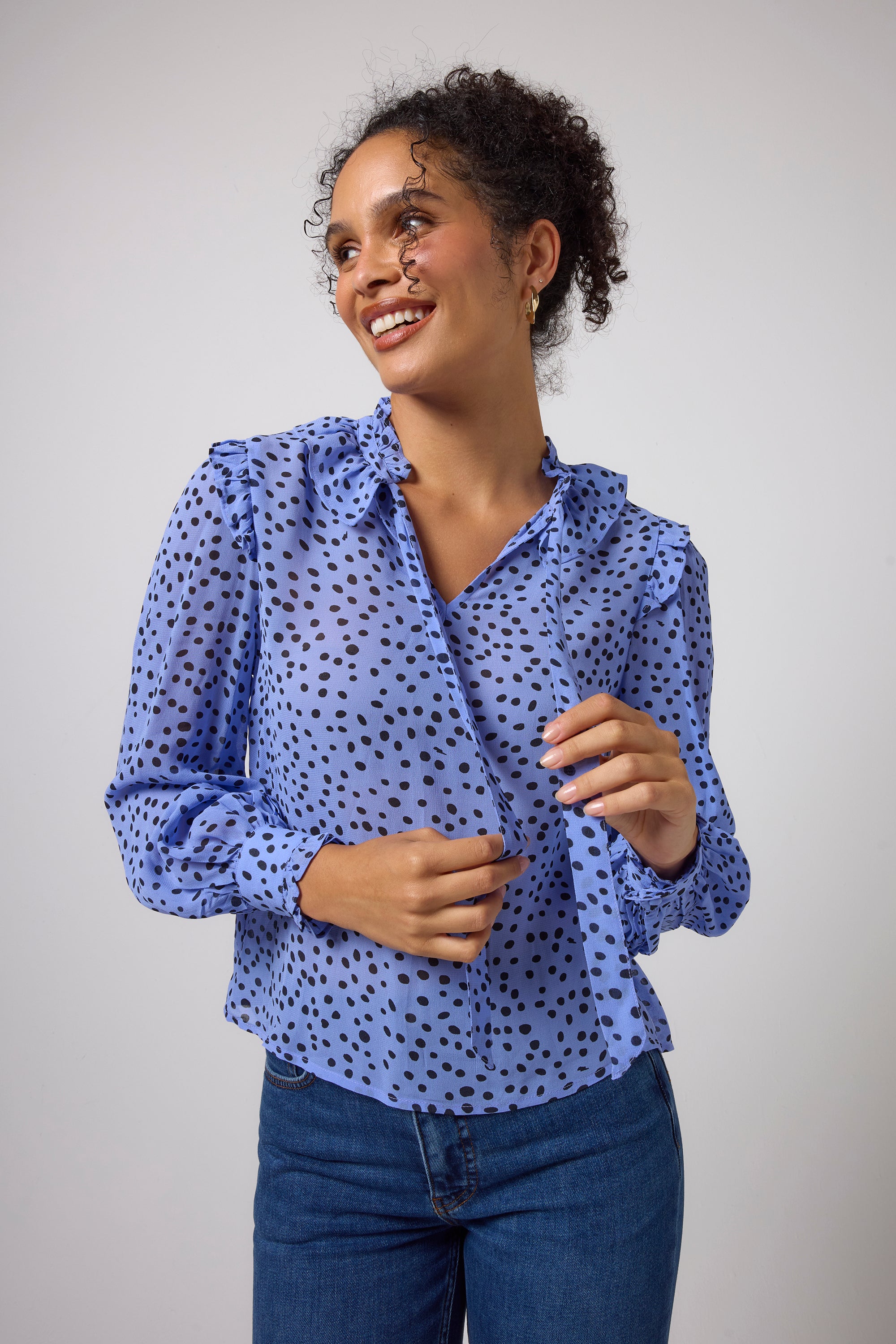 Scamp & Dude Blue with Black Cheetah Spot Tie Neck Blouse | Model wearing a light blue long-sleeve blouse with black polka dots and dark blue jeans.