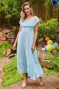 A blonde lady wearing a light blue with gold lurex spots Bardot midi dress on the shoulder with gold strappy heels