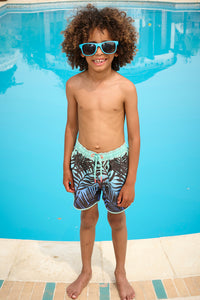 A curly-haired boy poolside wearing green with blue and black tropical and lightning bolt print swim shorts