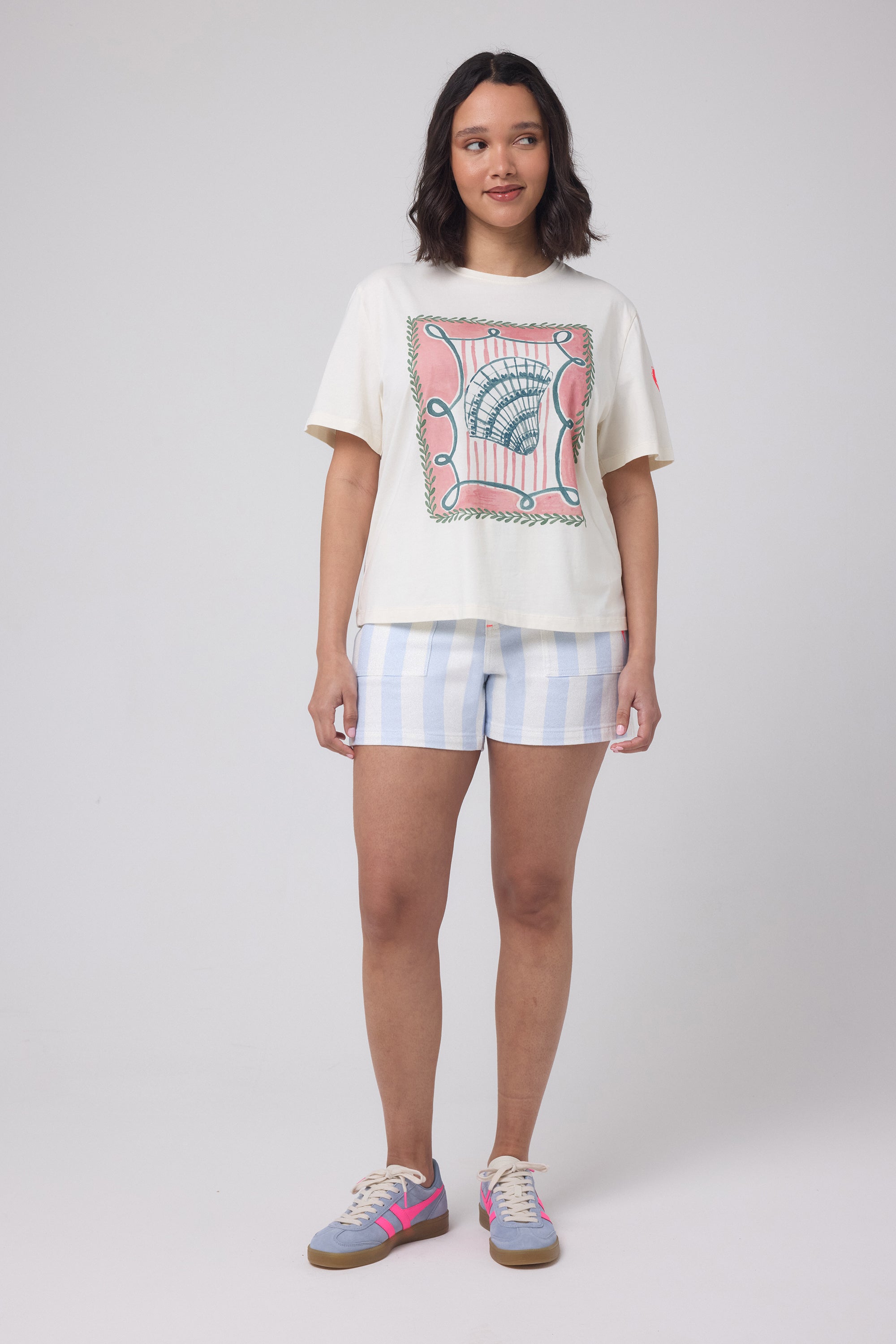 Scamp and Dude Cream Summer Shell Print T-Shirt | Model wearing a cream t-shirt with a shell graphic print. Worn with blue stripe denim shorts and trainers.