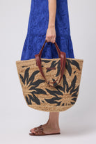 Scamp and Dude Natural with Black Sun Straw Basket Bag | Model wearing a blue broderie dress holding a straw bag with black sun design on it.