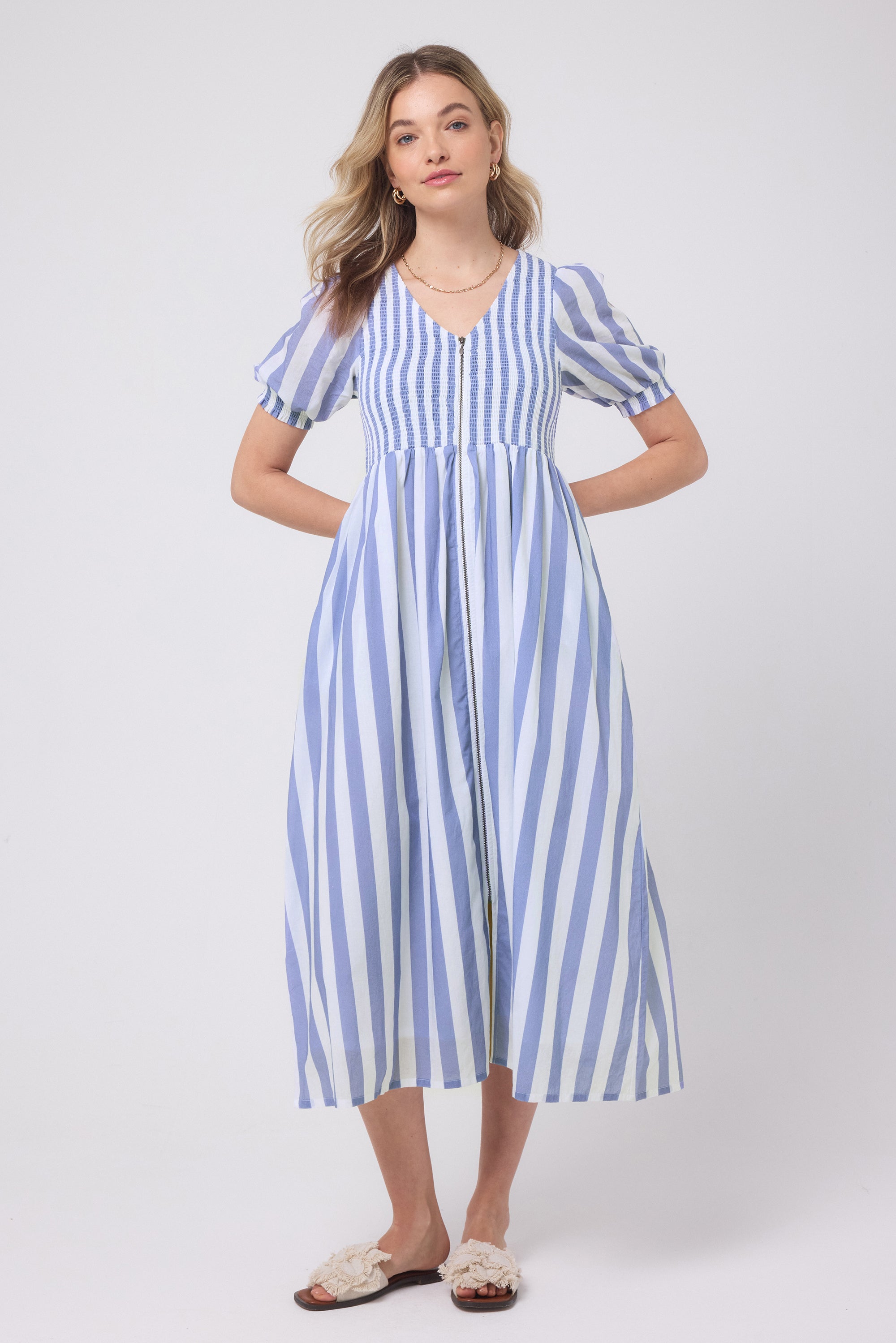 Scamp and Dude Blue with White Stripe Shirred Zip Detail Midi Dress | Model wearing a blue and white stripe zip front dress with neutral sandals and gold jewellery.
