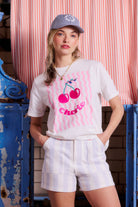 Scamp and Dude White Cherry Print T-Shirt | Model wearing a white t-shirt with a cherry print and a pair of stripe denim shorts.