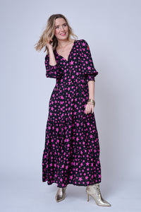 A blonde lady swishing the tiered skirt of her black maxi dress with magenta star and lightning bolt print, the dress has 3/4 length blouson sleeves, a tie V-neck and a neon pink embroidered lightning bolt on the sleeve