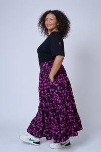 A lady wearing a black with magenta and blue snow leopard and star print tiered maxi skirt with a black t-shirt and trainers