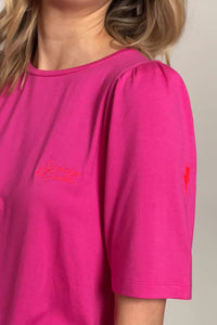 A close-up pink T-shirt with embroidered neon coral Scamp & Dude handwritten logo on the chest and neon pink embroidered lightning bolt on the arm