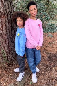 Two boys running in the woods wearing matching sweatshirts in different colourways, one is wearing pink with a neon pink towelling balloon on the chest, the other is wearing blue with a yellow balloon