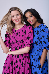 Two women wearing matching star & lightning bolt dresses in different colourways, one is magenta, the other is blue