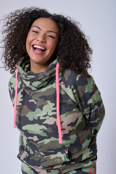 A curly-haired lady wearing a khaki camo cowl neck with neon coral cords & a neon pink embroidered lightning bolt on the arm