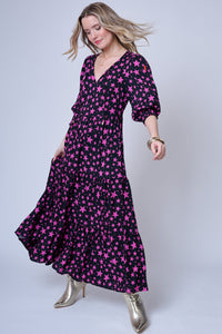 A blonde lady wearing a black maxi dress with magenta star and lightning bolt print with gold heeled boots, the dress has 3/4 length blouson sleeves, a tiered skirt and a neon pink embroidered lightning bolt on the sleeve