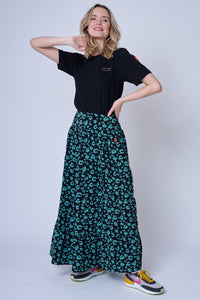 A blonde lady wearing a khaki leopard and lightning bolt print tiered maxi skirt with a neon pink embroidered lightning bolt on the hip, a tucked in black t-shirt and colourful trainers