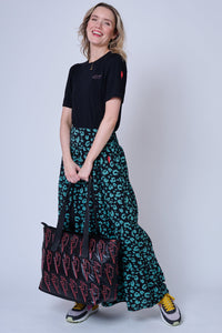 A blonde lady wearing a khaki leopard and lightning bolt print tiered maxi skirt with a neon pink embroidered lightning bolt on the hip, a tucked in black t-shirt and colourful trainers, she is holding a black overnight bag with coral lightning bolts on