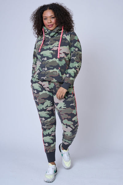 A curly-haired lady wearing khaki camo print joggers with a matching cowl neck hoodie and nike trainers