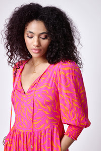 A curly-haired lady wearing a pink with orange zebra and lightning bolt print tie front maxi dress with 3/4 blouson sleeves