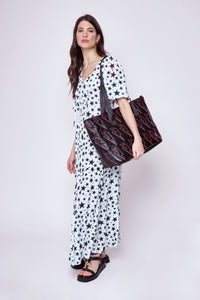A lady wearing an ivory with black star & lightning bolt print tie front tiered maxi dress holding a black metallic bag