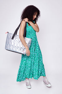 A lady holding a silver overnight bag wearing a bright green and lilac zebra and lightning bolt print maxi sundress