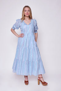A blonde lady wearing a pale blue tie front maxi dress with rainbow lurex stripes with a tiered skirt and 3/4 blouson sleeves