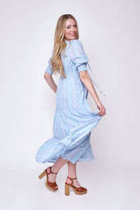 A blonde lady swishing the skirt of her pale blue tie front maxi dress with rainbow lurex stripes