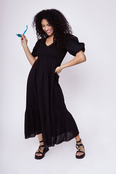 A curly-haired lady wearing a black v-neck shirred dobby maxi dress with puff sleeves and black sandals