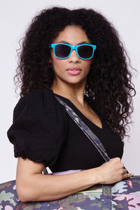 A curly-haired lady wearing neon blue sunglasses with black lightning bolts on the front and smoke coloured lenses