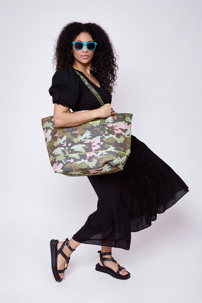 A lady wearing a black dobby Scamp & Dude dress holding a khaki camo print with neon pink outlines weekender bag