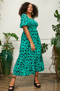 A curly-haired lady wearing a green with black leopard and lightning bolt print shirred midi dress with blouson sleeves