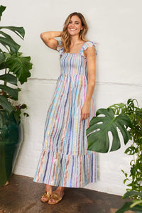 A blonde lady wearing a pale blue with rainbow lurex stripe detail maxi sundress with gold sandals