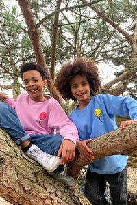 Two boys climbing a tree wearing matching sweatshirts in different colourways, one is wearing pink with a neon pink towelling balloon on the chest, the other is wearing blue with a yellow balloon
