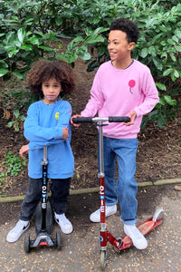 Two boys with scooters wearing matching sweatshirts in different colourways, one is wearing pink with a neon pink towelling balloon on the chest, the other is wearing blue with a yellow balloon