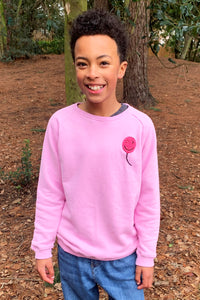A boy wearing a pink sweatshirt with a neon pink towelling balloon on the chest and blue jeans