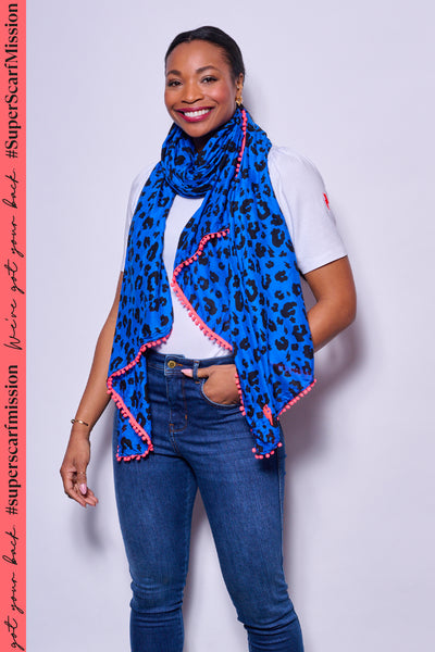 A lady wearing a blue with black leopard & lightning bolt print scarf with a neon pink pom pom trim with jeans and a t-shirt