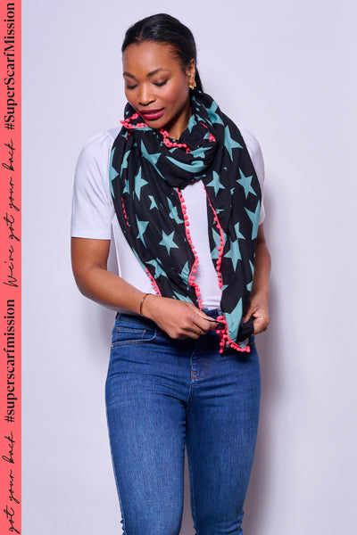 A lady wearing a black with khaki star print scarf featuring a neon pink pom pom trim with a white t-shirt and blue jeans